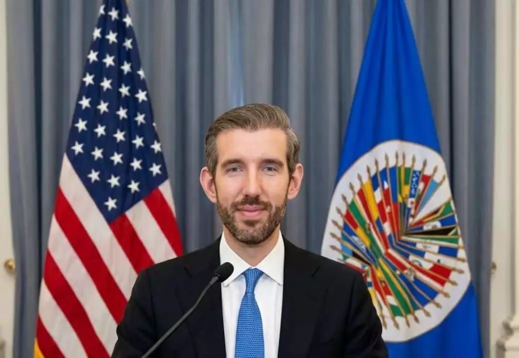 Homem de terno escuro e gravata azul fala ao microfone diante das bandeiras dos Estados Unidos e da Organização dos Estados Americanos