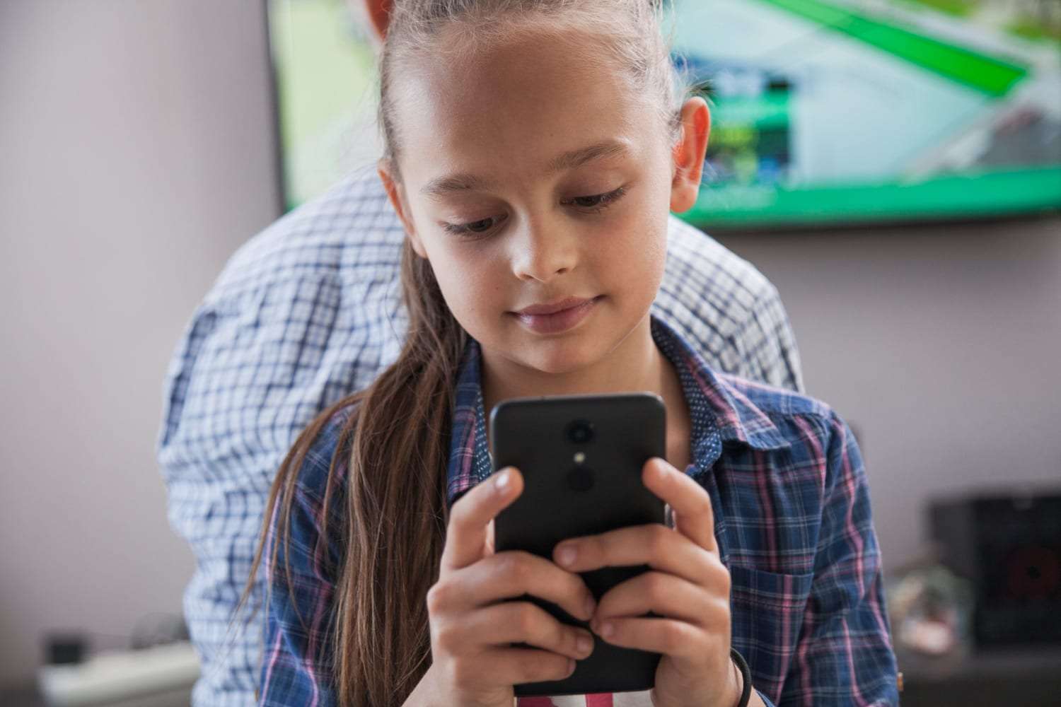 Menina de camisa xadrez segura um celular com as duas mãos e olha para a tela, com um adulto desfocado ao fundo