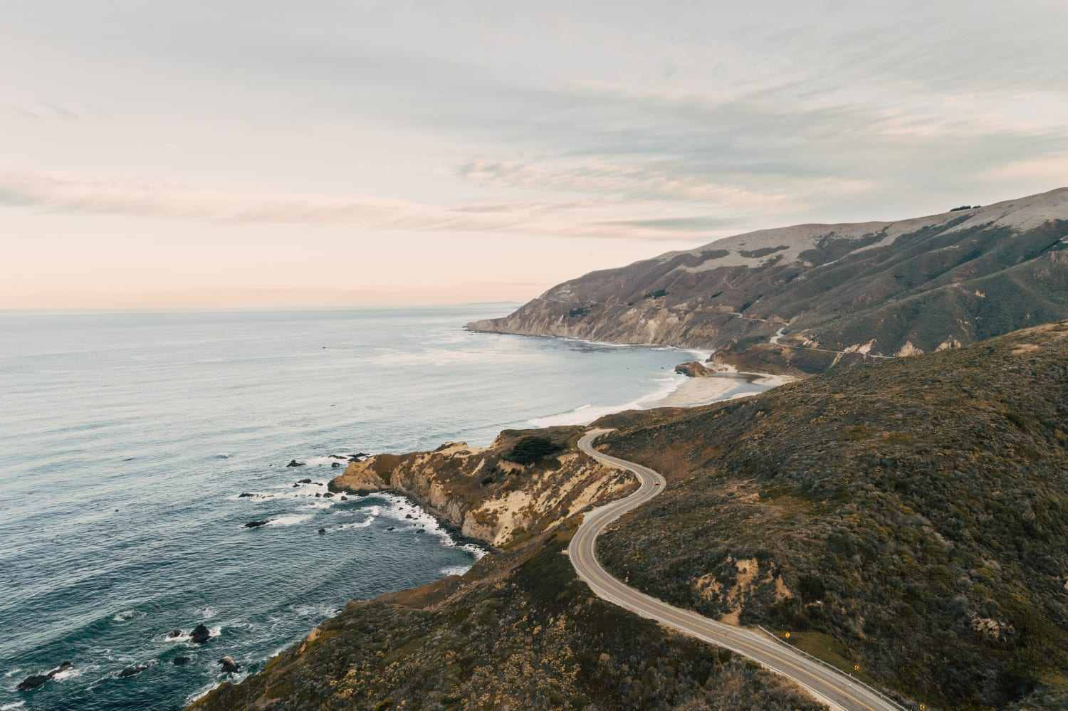 Vista aérea de uma estrada de duas pistas que serpenteia por um morro à beira do oceano, com falésias e céu alaranjado ao entardecer