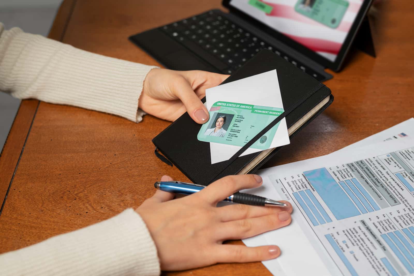 Mão segurando um green card sobre um envelope, com formulário de imigração e tablet ao fundo
