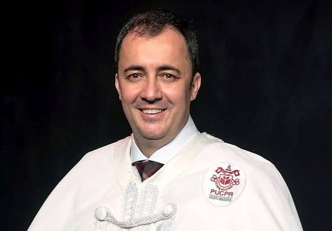 Homem de cabelo curto, sorrindo, vestido com beca branca com o emblema da PUCPR, diante de fundo preto