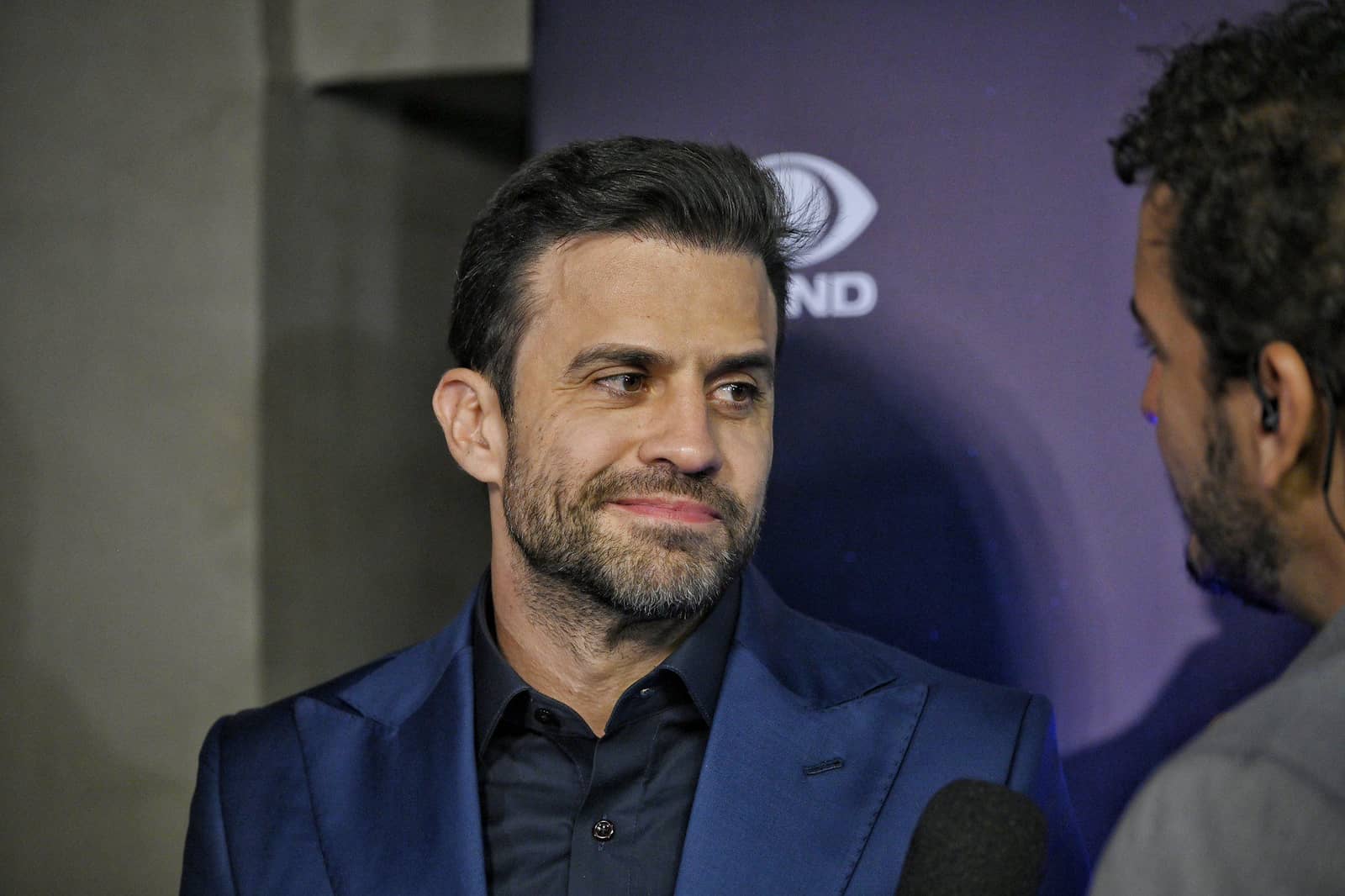 Homem de barba, paletó azul e camisa preta olha para o lado durante entrevista, diante de um painel roxo com o logotipo de uma emissora de televisão