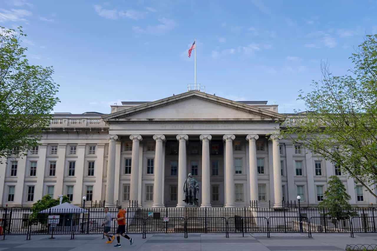 Fachada com colunas do prédio do Departamento do Tesouro dos Estados Unidos, em Washington, com bandeira americana no mastro e dois corredores passando na calçada
