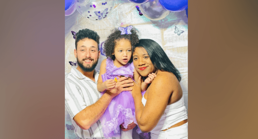 Casal segura no colo uma menina pequena de vestido lilás em frente a um painel de festa com balões roxos e borboletas de papel