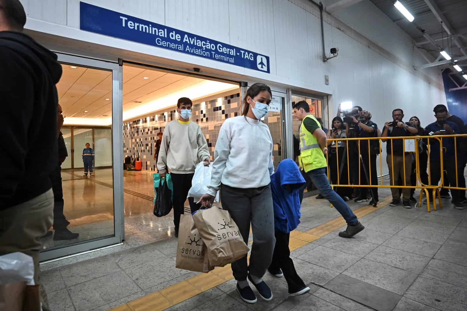 Passageiros de máscara carregam sacolas ao sair de um terminal de aviação geral, com jornalistas atrás de grades amarelas registrando a chegada