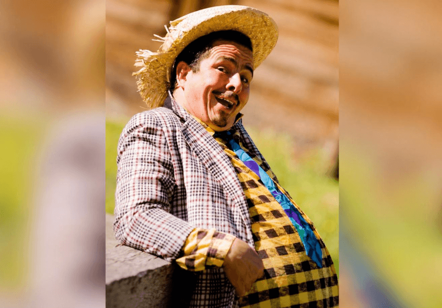 Homem de chapéu de palha, paletó xadrez e gravata colorida faz careta apoiado em um muro, caracterizado como personagem caipira
