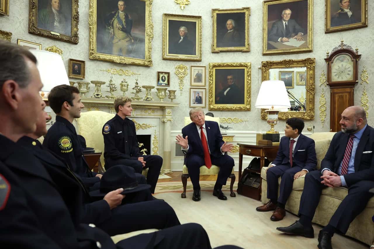 Reunião no Salão Oval da Casa Branca, com salva-vidas de uniforme escuro sentados de um lado e, do outro, um menino de terno ao lado de um adulto