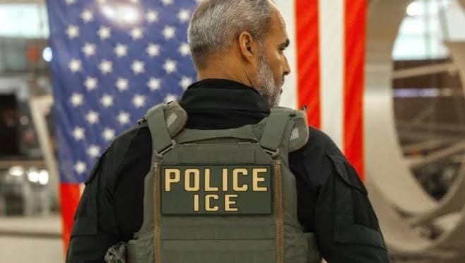 Agente de costas, com colete verde escrito Police ICE, diante de uma grande bandeira dos EUA em um saguão