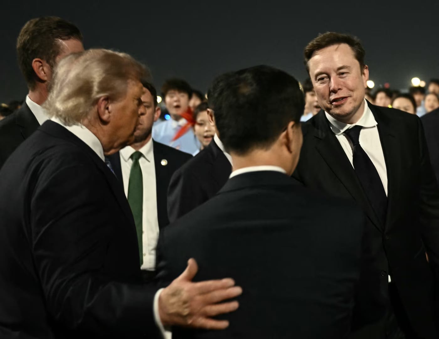 Elon Musk cumprimenta autoridades em pista de aeroporto à noite, ao lado do presidente Donald Trump, com público atrás das autoridades