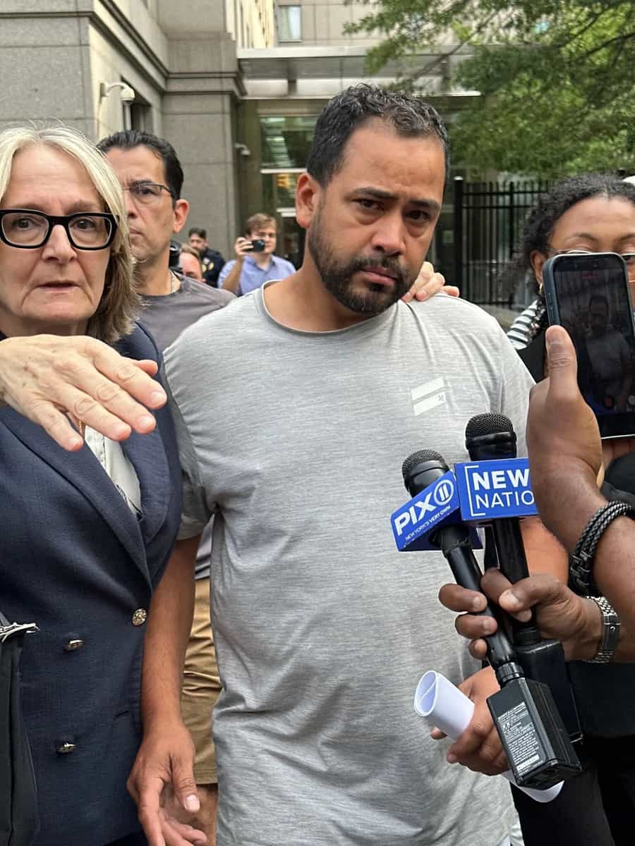 Homem de camiseta cinza deixa prédio do tribunal federal cercado por microfones de emissoras de televisão e por advogada de terno escuro