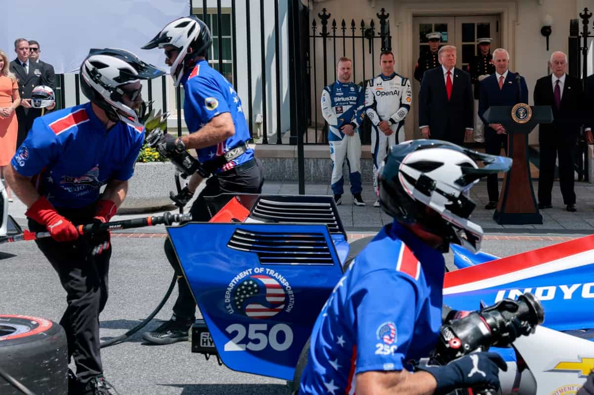 Equipe de boxes com uniforme azul e capacete branco simula troca de pneus de carro de IndyCar em frente à Casa Branca, com pilotos e autoridades assistindo