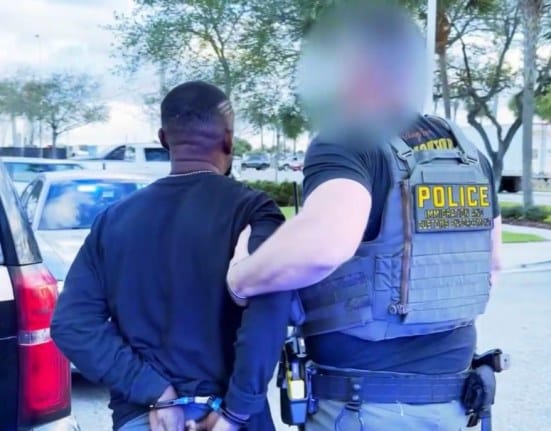 Homem algemado sendo conduzido por um agente com colete escrito POLICE, ao lado de veículos, em imagem ilustrativa de ação de imigração