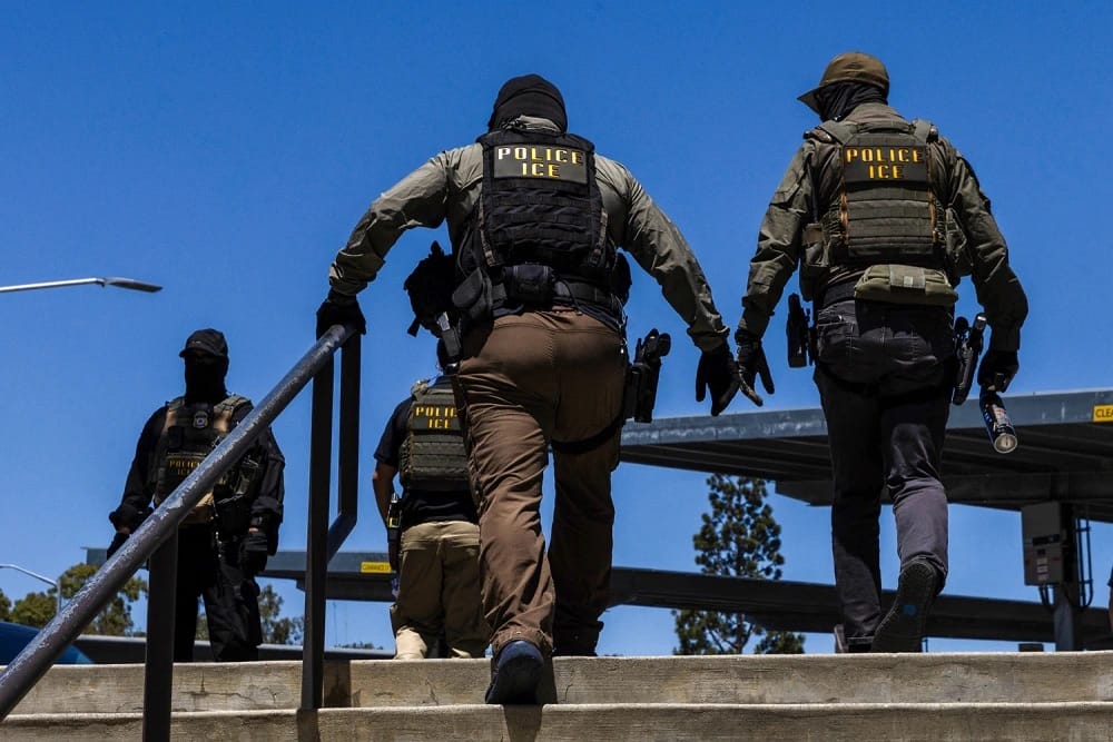 Quatro agentes com coletes escuros marcados POLICE ICE e rostos cobertos sobem uma escada de concreto sob céu azul