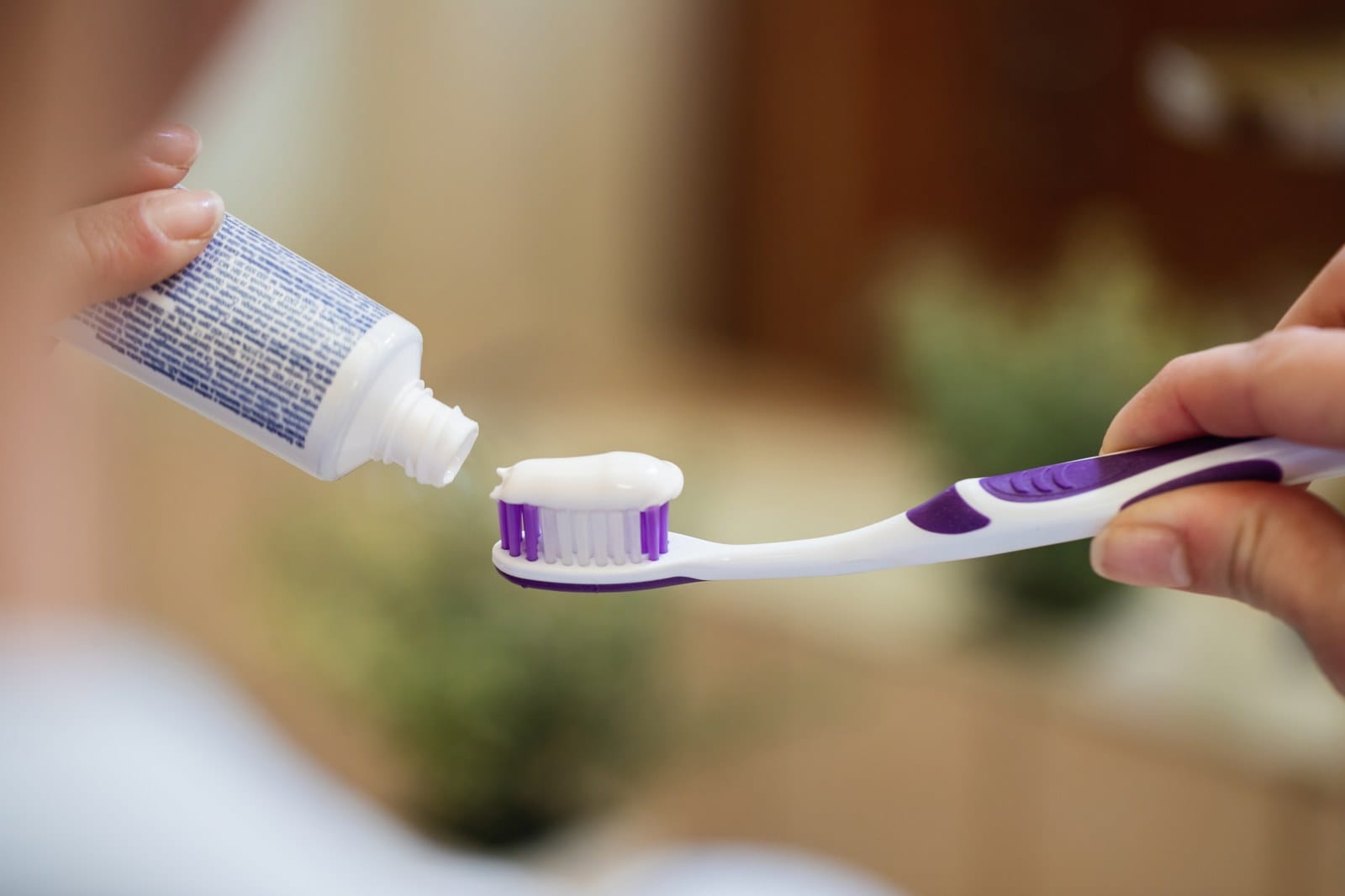 Mão aplica pasta de dente branca em uma escova roxa e branca, com o tubo aberto ao lado