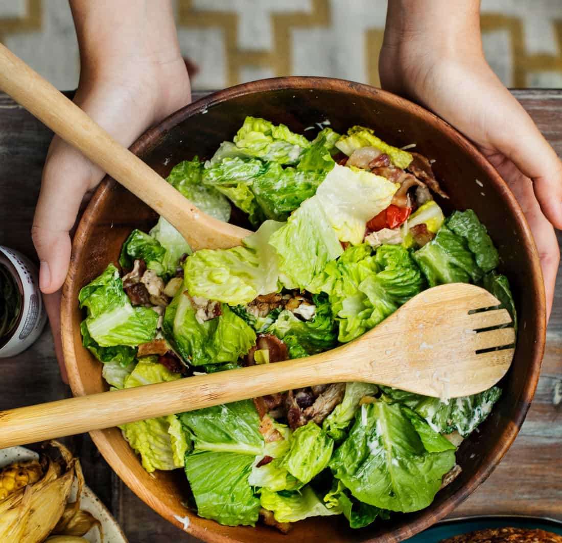 Tigela de madeira com salada de alface, tomate e outros ingredientes, com duas colheres de madeira e mãos segurando a tigela