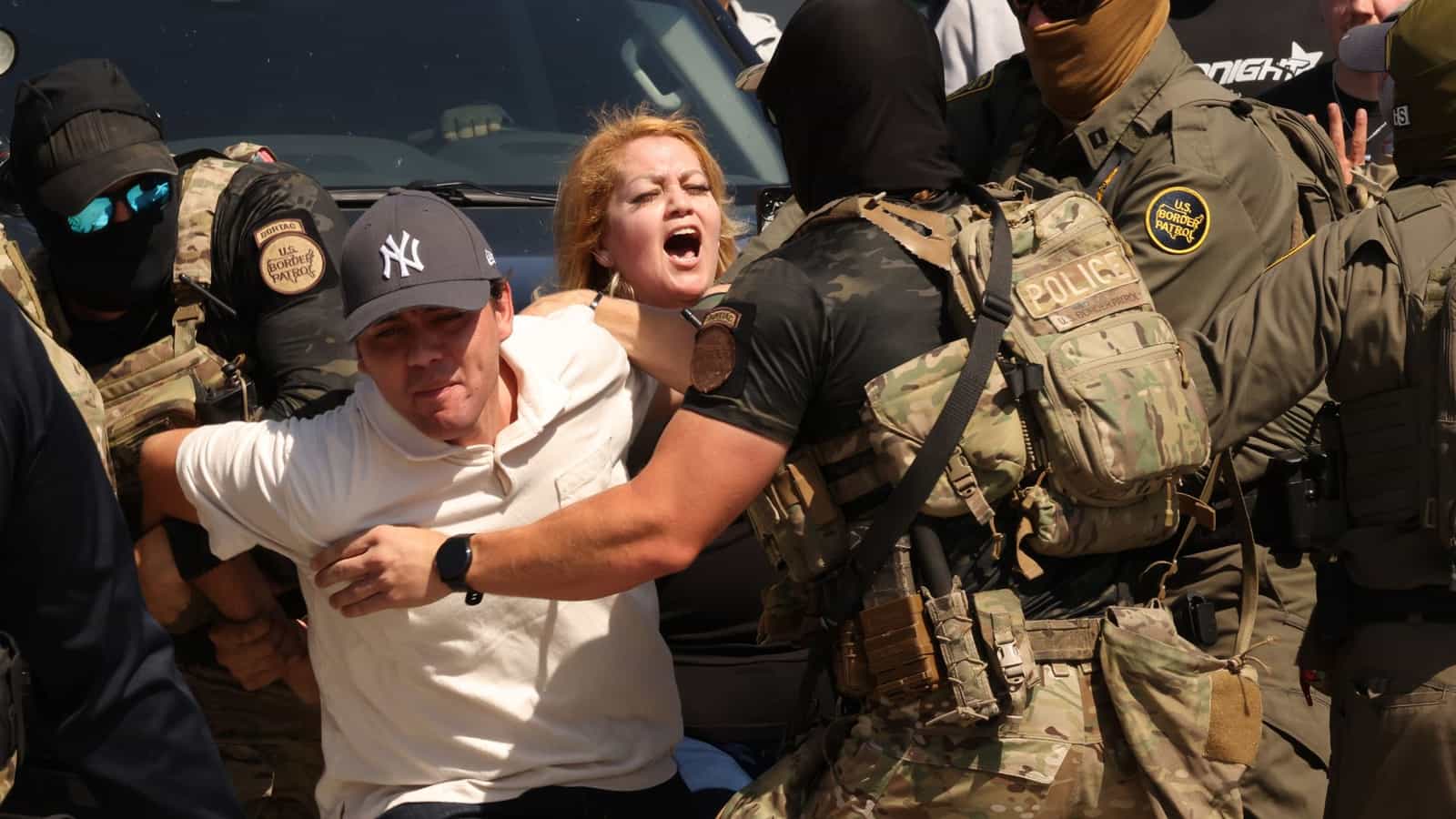 Agentes mascarados com coletes da U.S. Border Patrol e da Polícia imobilizam um homem de camiseta branca enquanto uma mulher grita ao lado, diante de um veículo
