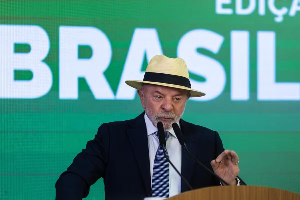 O presidente Lula, de terno escuro e chapéu de palha, fala ao microfone diante de um painel verde com a palavra BRASIL