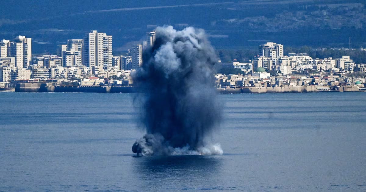 Coluna de fumaça escura sobe da água diante de uma cidade litorânea no Oriente Médio, com prédios ao fundo