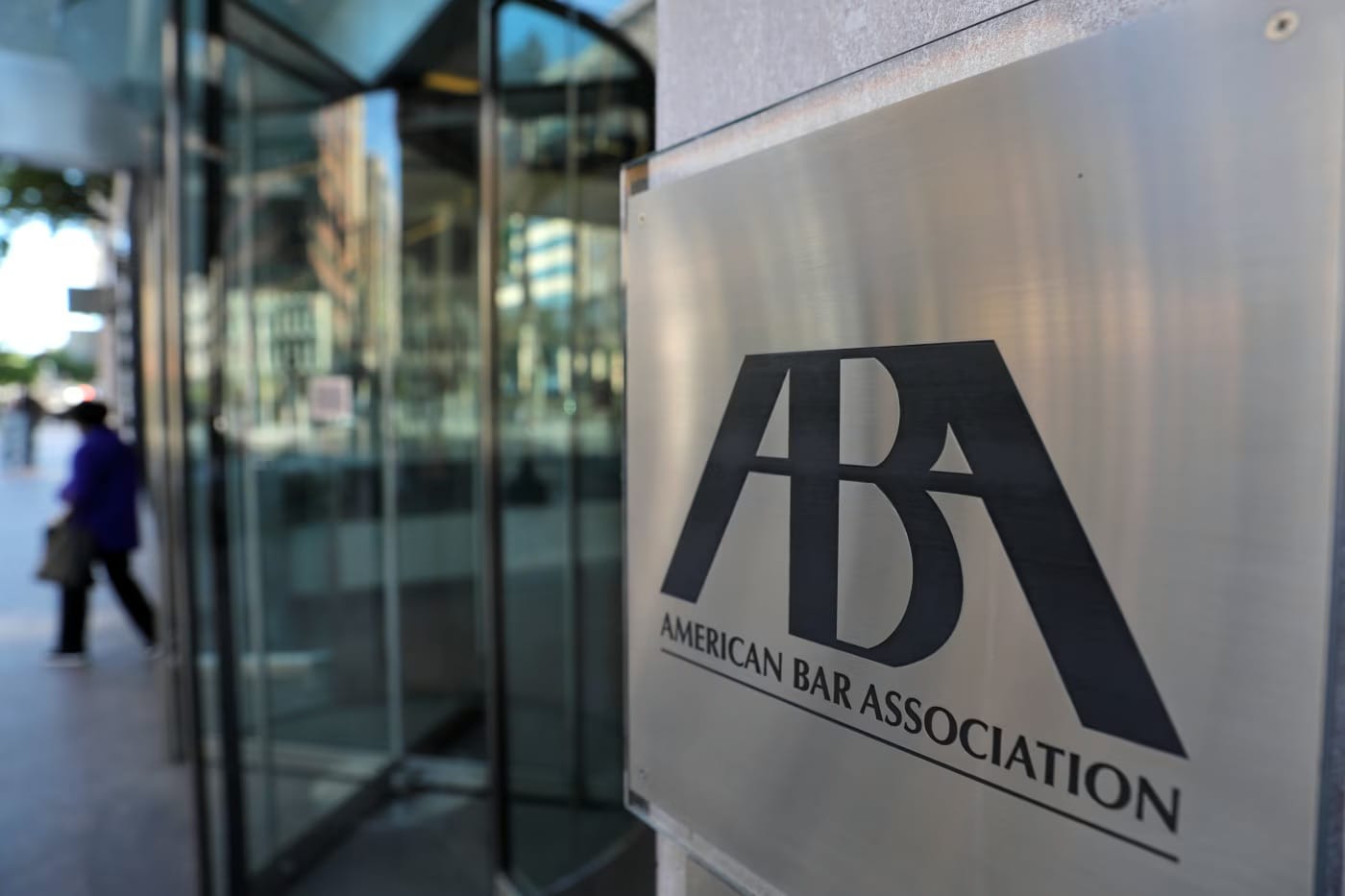 Placa metálica com o logotipo e o nome American Bar Association na fachada de um prédio de vidro, com porta giratória ao lado