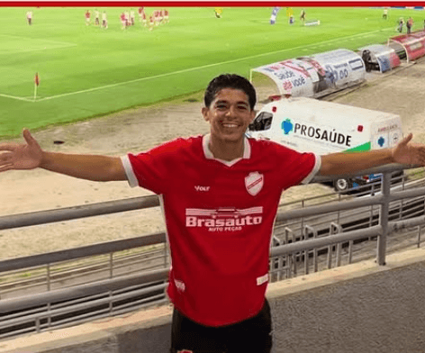 Homem sorridente com os braços abertos, vestindo camisa vermelha de time de futebol, em um estádio