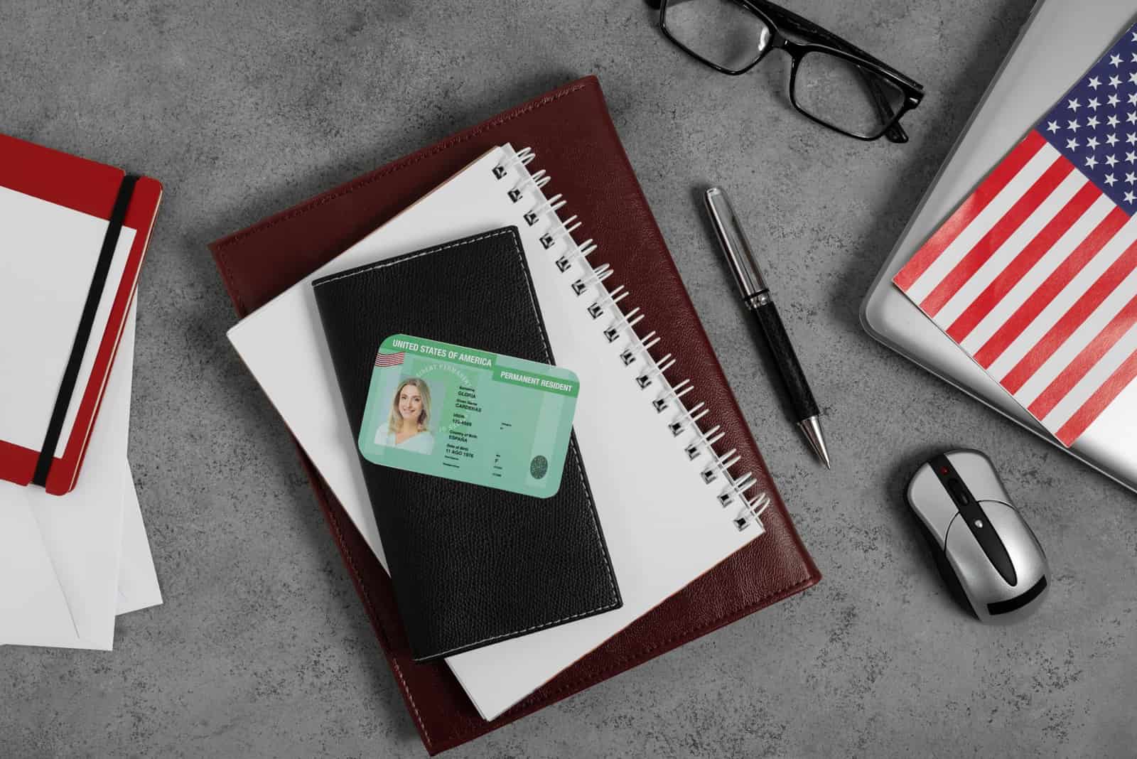 Green card sobre uma agenda, ao lado de óculos, caneta e bandeira dos Estados Unidos