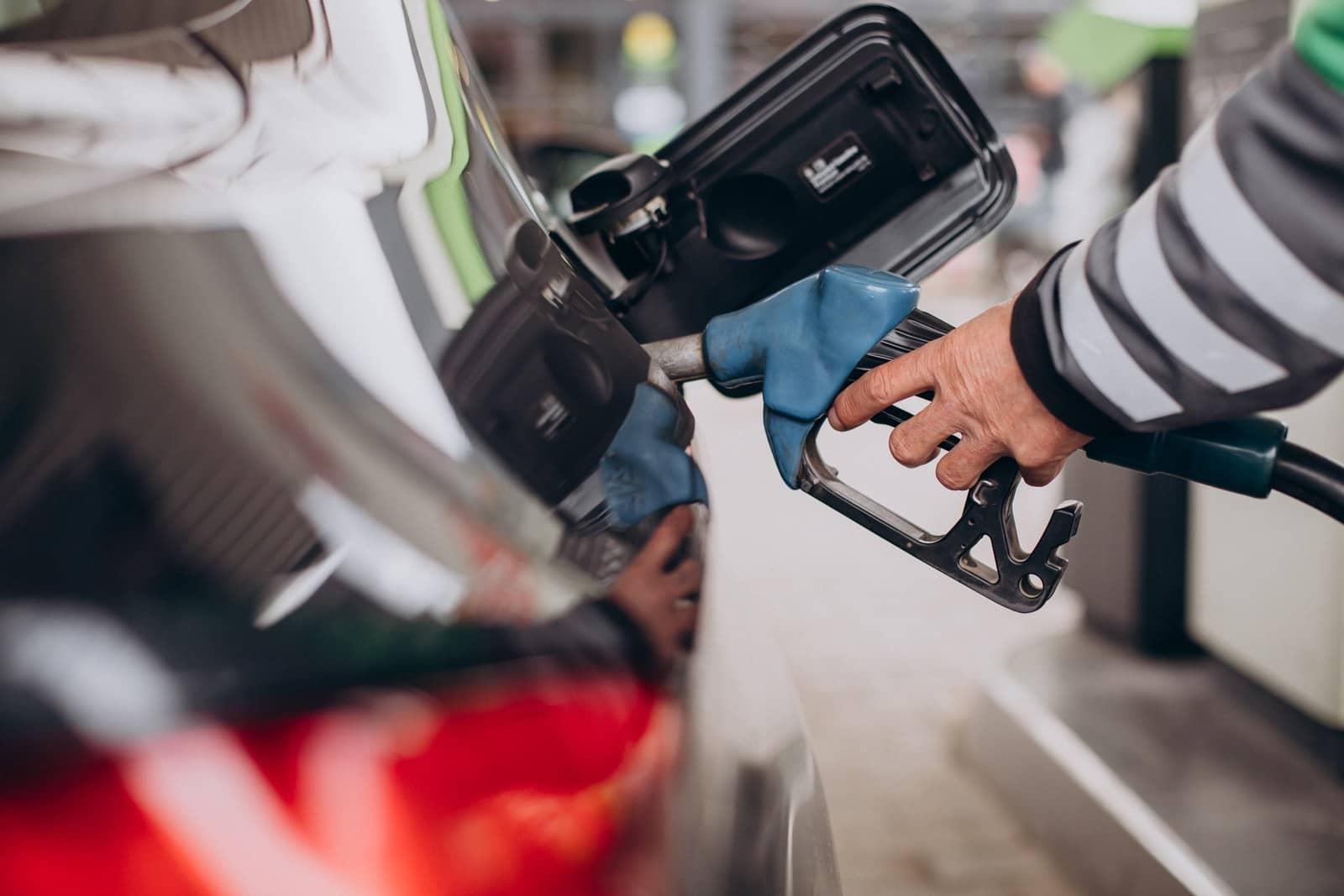 Mão com luva de listras cinza segura a bomba de combustível azul no tanque de um carro, em um posto de gasolina