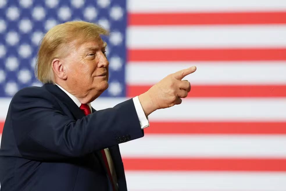 Donald Trump, de terno escuro e gravata vermelha, aponta com a mão direita diante de uma bandeira dos Estados Unidos