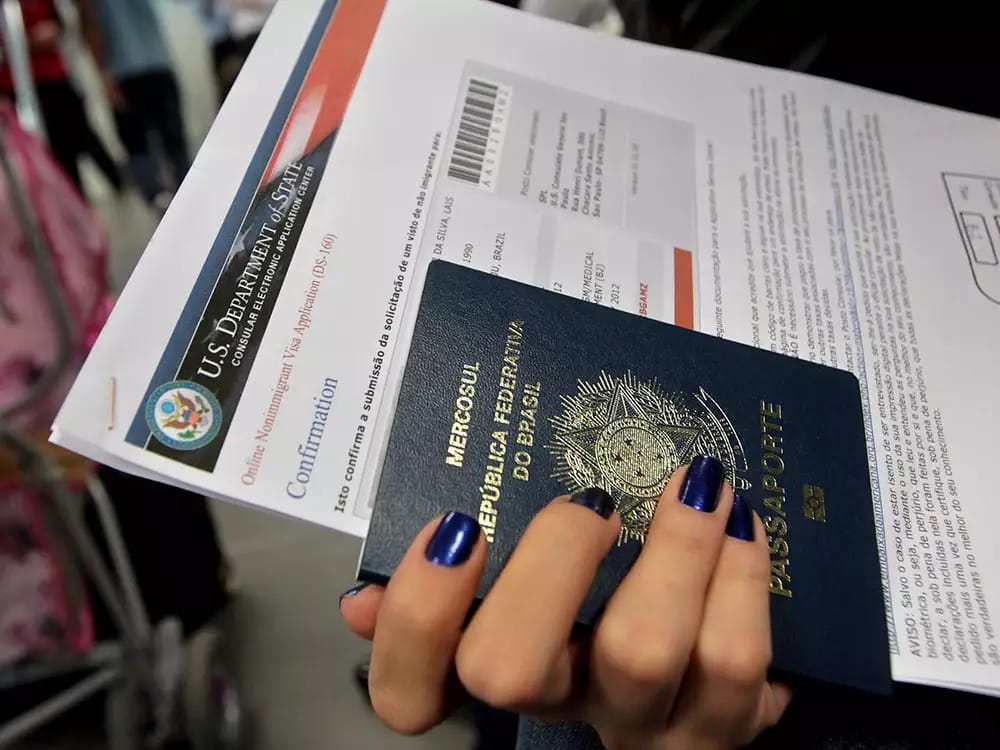 Mão segura um passaporte brasileiro e um formulário de solicitação de visto americano