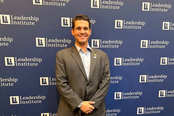 Christopher Costa, de terno, posa diante de um painel com o logotipo do Leadership Institute