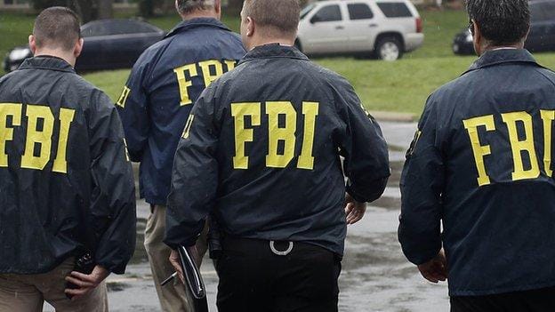Quatro agentes do FBI, vistos de costas, com jaquetas escuras estampadas com as letras FBI em amarelo, caminham por um estacionamento