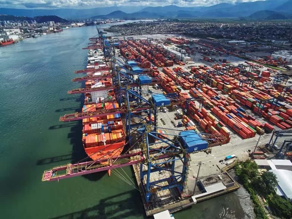 Vista aérea do Porto de Santos com navio cargueiro atracado e milhares de contêineres coloridos empilhados sob guindastes