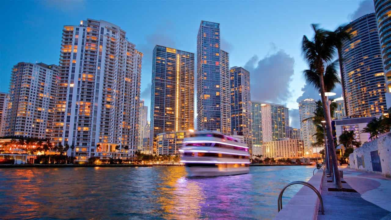 Skyline de arranha-céus iluminados de Brickell, em Miami, ao entardecer, com um barco deixando rastro de luz na água e uma palmeira à direita