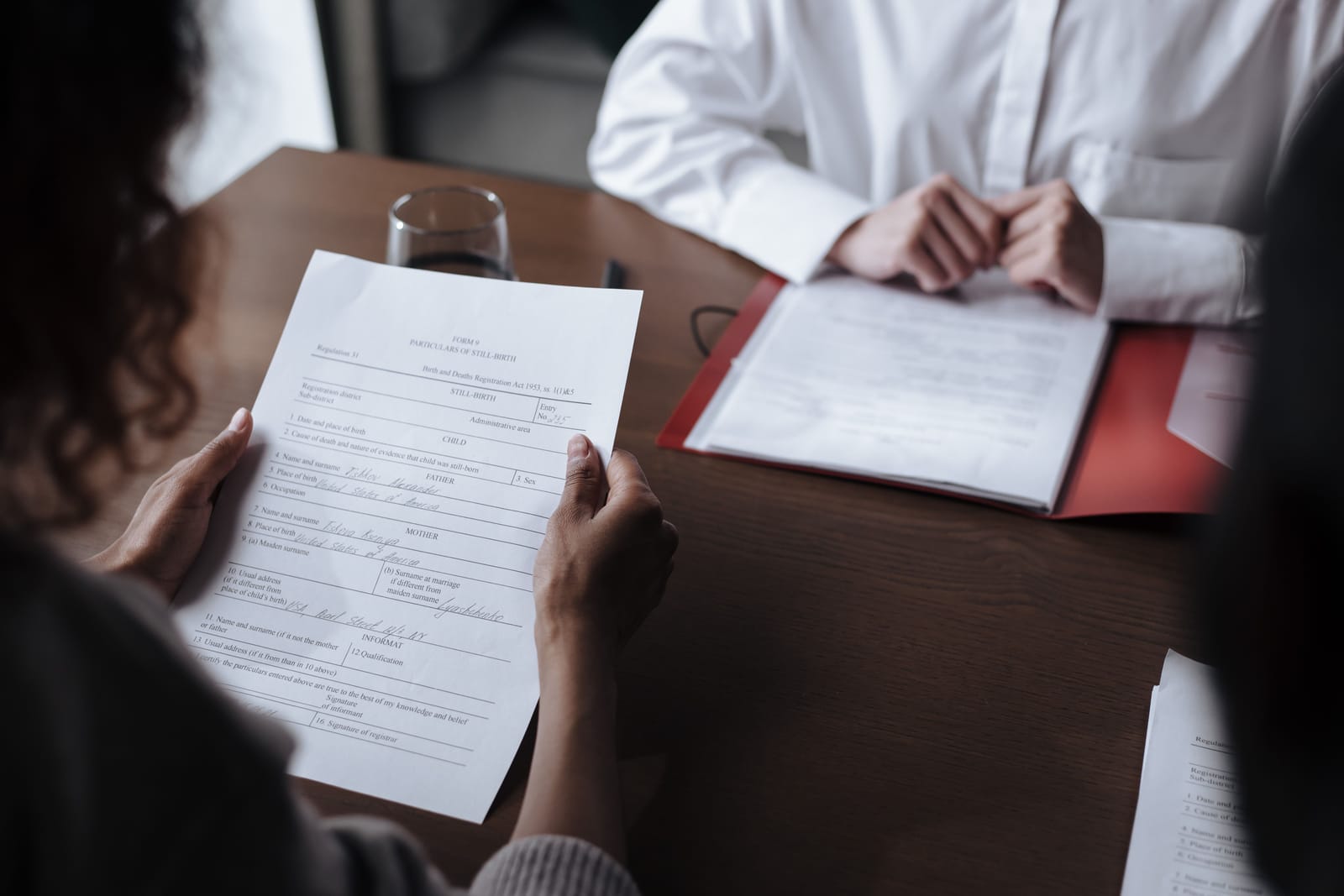 Duas pessoas sentadas à mesa de escritório durante um atendimento, uma delas segurando um formulário impresso enquanto a outra folheia documentos em uma pasta vermelha