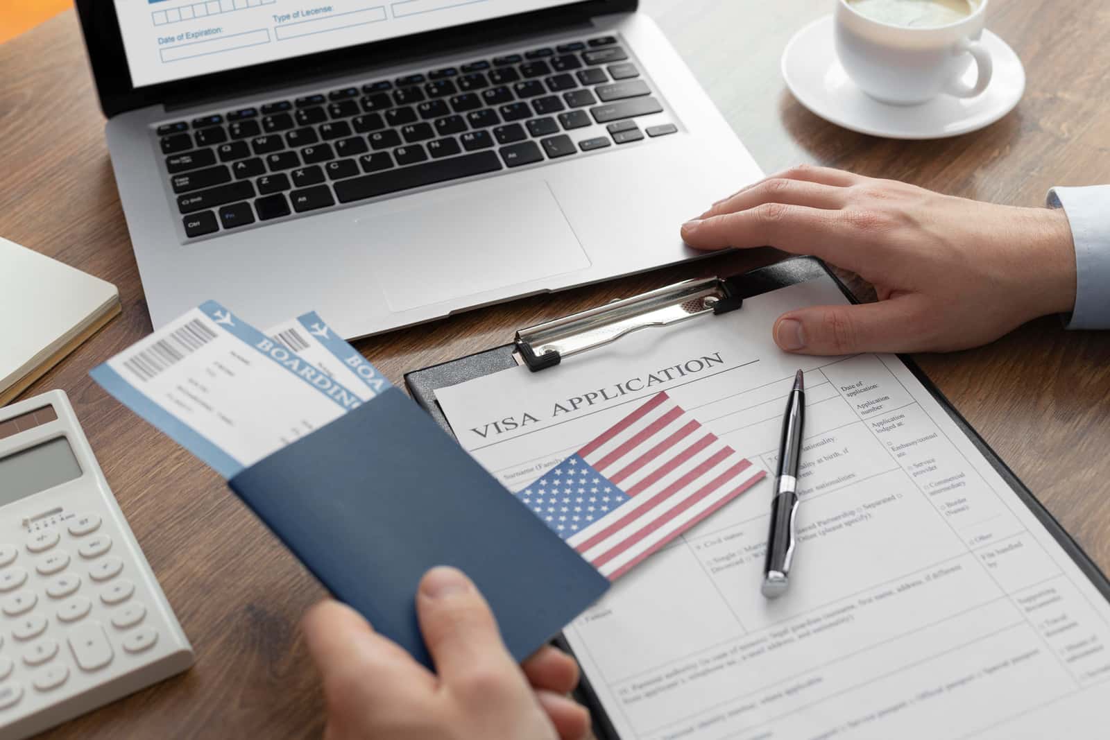 Formulário de solicitação de visto americano sobre prancheta, ao lado de passaporte com cartões de embarque, bandeira dos Estados Unidos, caneta e calculadora