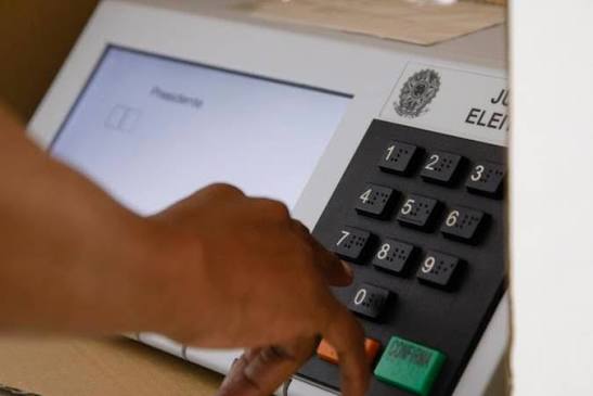 Mão pressiona o teclado numérico de uma urna eletrônica brasileira, com a tela mostrando o cargo de presidente