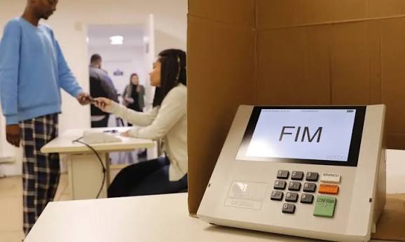Urna eletrônica exibindo a mensagem FIM ao lado de uma mesária que entrega o documento a um eleitor