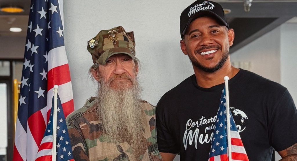 Barbeiro brasileiro Diego Henrique, com camiseta e boné do projeto Cortando pelo Mundo, ao lado de veterano americano fardado entre bandeiras dos EUA