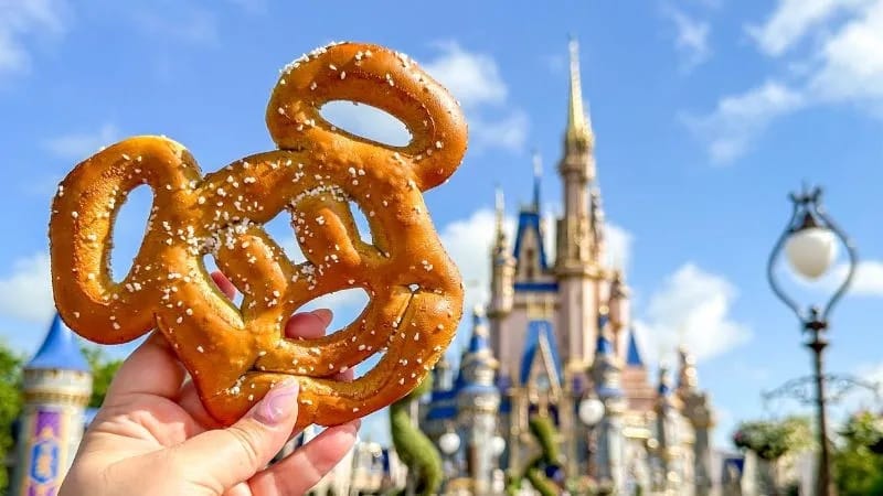 Mão segura um pretzel salgado em formato da cabeça do Mickey Mouse tendo ao fundo o castelo de um parque da Disney sob céu azul