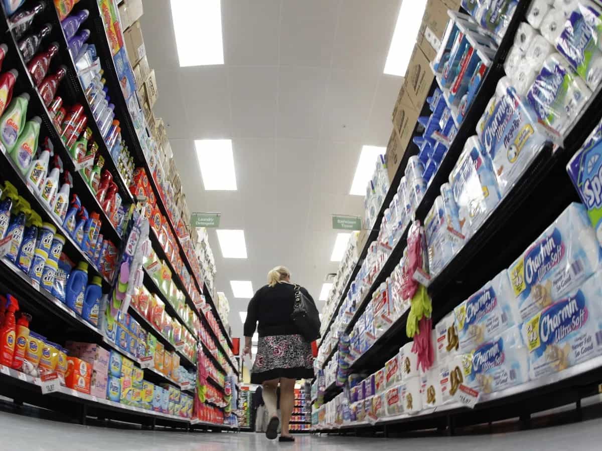 Mulher de costas caminha por um corredor de supermercado com prateleiras repletas de produtos de limpeza e pacotes de papel higiênico dos dois lados