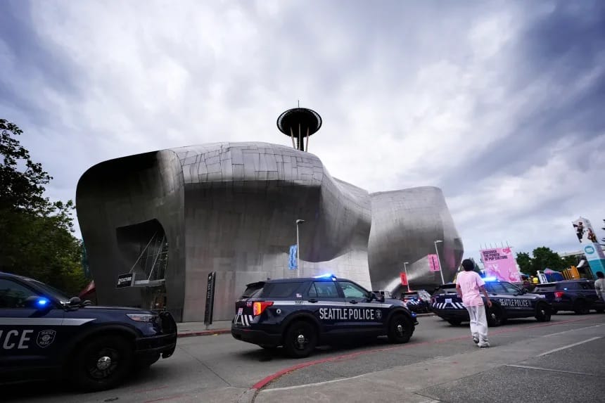 Viaturas da polícia de Seattle estacionadas em frente ao prédio metálico do museu MoPOP, com a Space Needle ao fundo sob um céu nublado