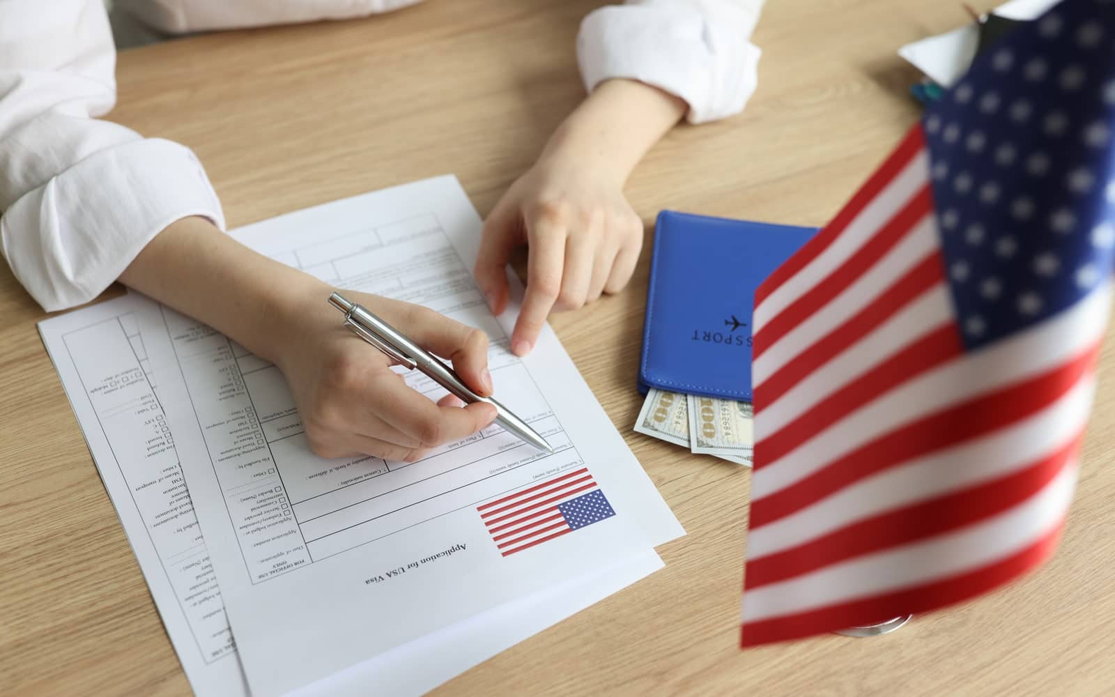 Mão preenchendo formulário de aplicação para visto dos Estados Unidos, ao lado de passaporte, dinheiro e bandeira americana