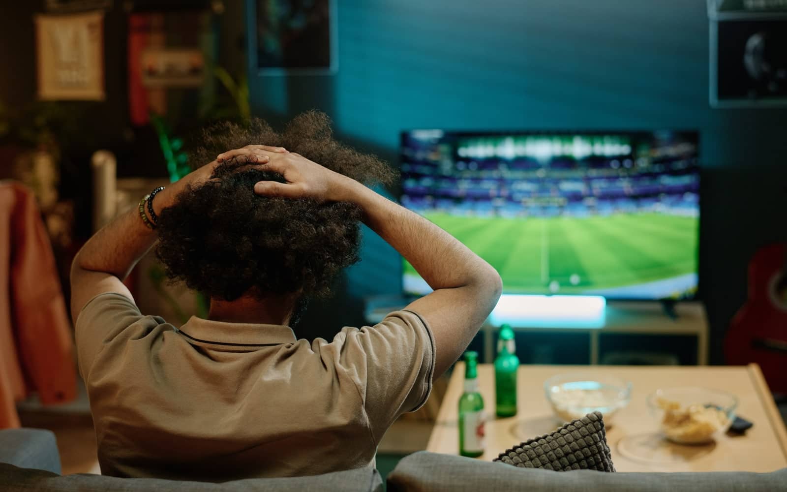 Homem visto de costas, com as mãos na cabeça, assistindo a uma partida de futebol na televisão em ambiente escuro