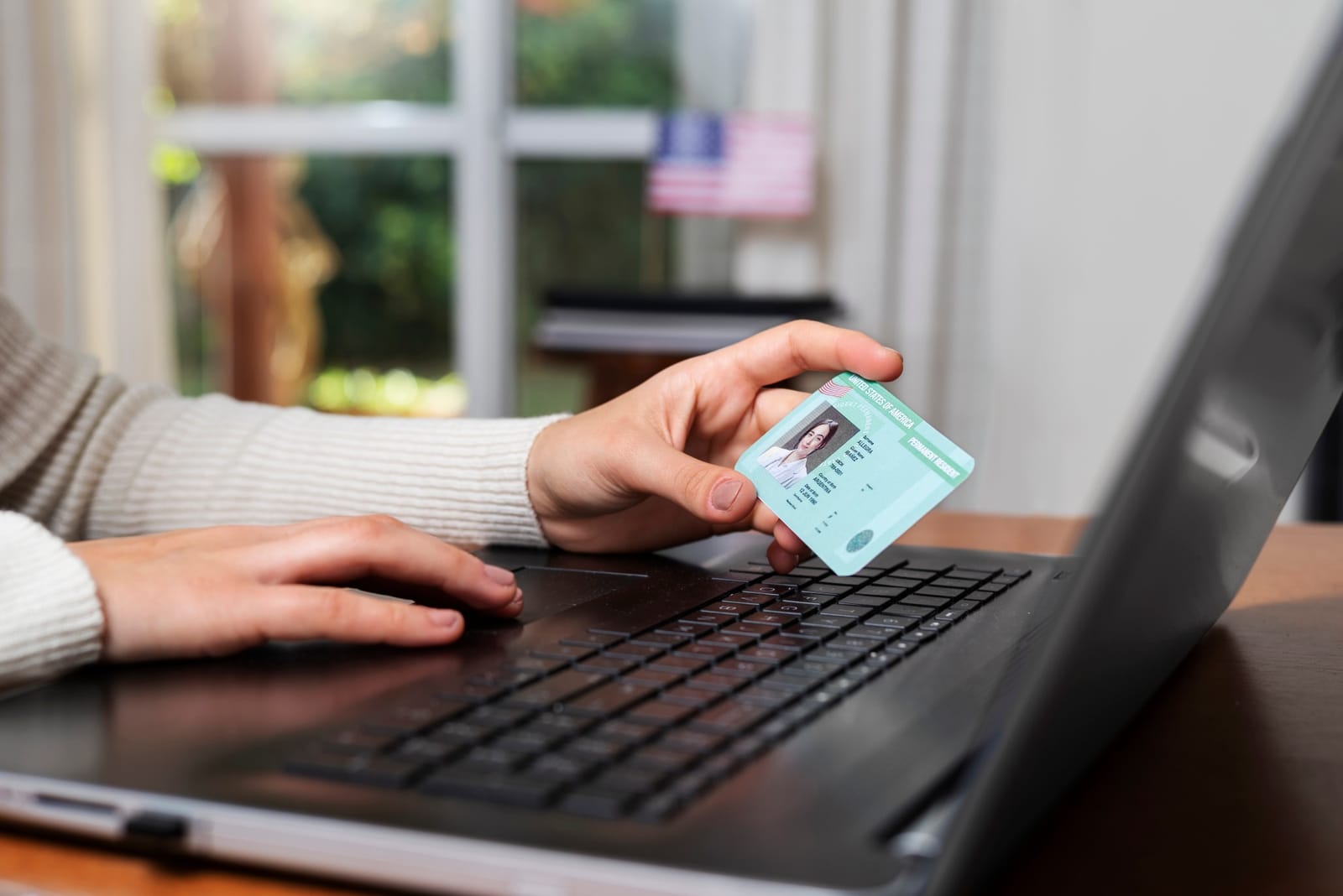 Mão segurando um green card sobre o teclado de um notebook, com bandeira dos Estados Unidos ao fundo