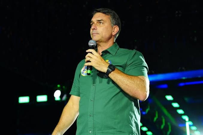 Flávio Bolsonaro, de camisa verde e relógio no pulso, fala ao microfone em um palco escuro com luzes ao fundo