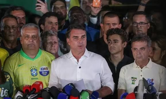 Flávio Bolsonaro, de camisa branca, fala diante de microfones e cercado por apoiadores, alguns de camisa da seleção brasileira