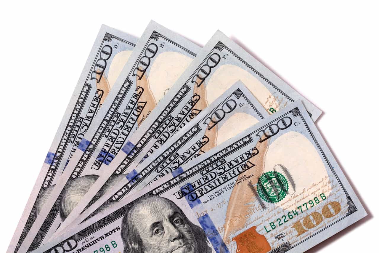 Cinco notas de 100 dólares abertas em leque sobre fundo branco, com o retrato de Benjamin Franklin em destaque