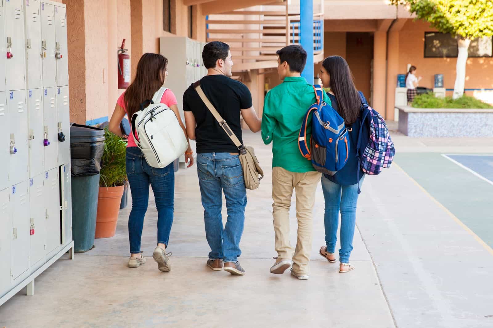 Quatro estudantes de costas, com mochilas, caminham juntos por um corredor externo de escola ao lado de armários