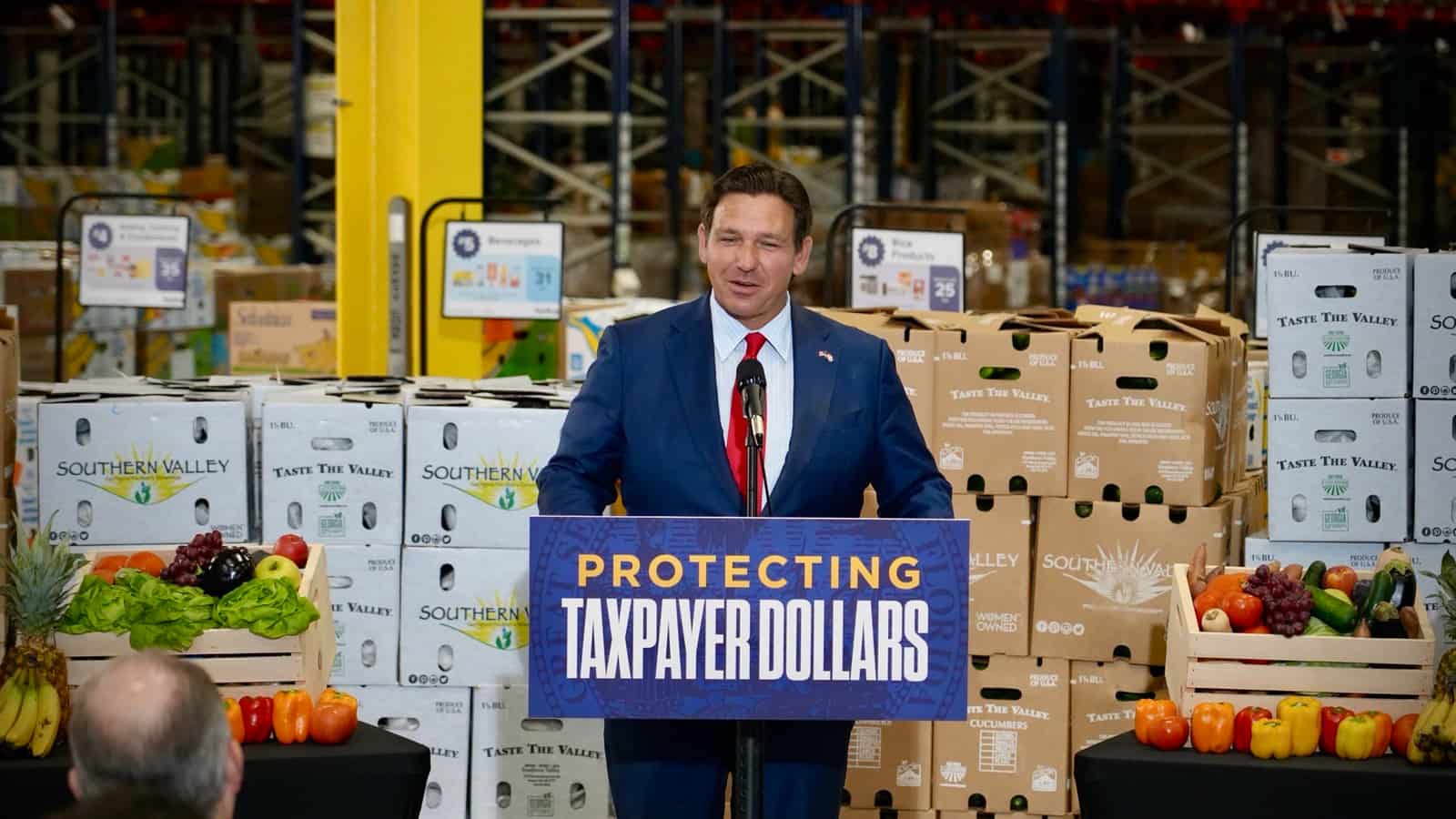 Governador da Flórida Ron DeSantis discursa em coletiva de imprensa atrás de púlpito com faixa Protecting Taxpayer Dollars