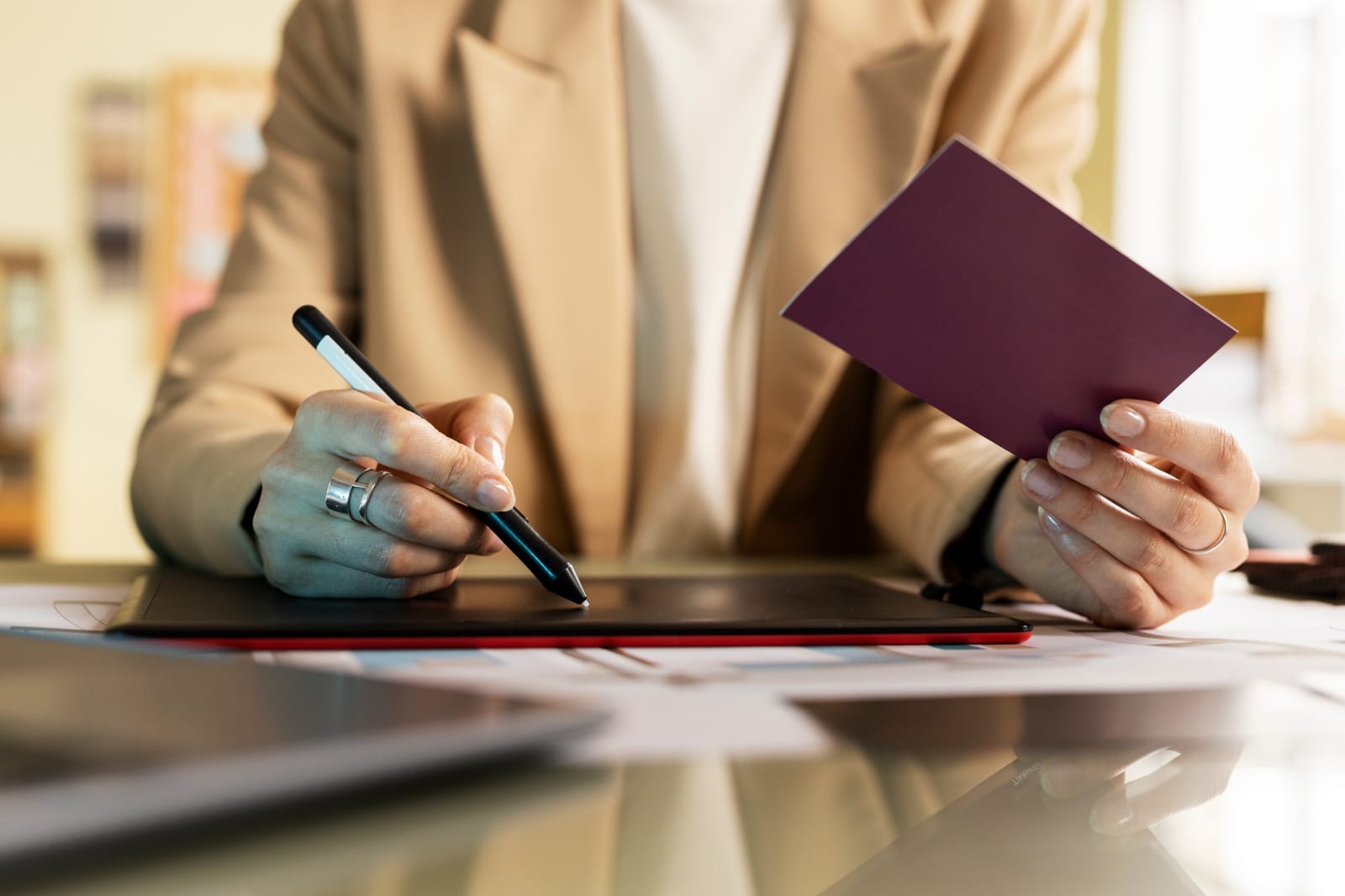 Mão segurando um passaporte, com outra mão usando caneta digital sobre uma mesa de escritório