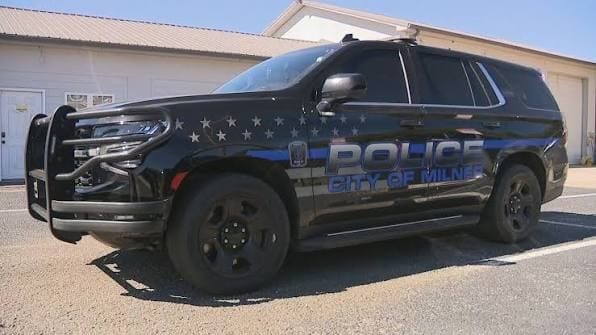 Viatura preta da polícia da cidade de Milner, com faixa azul e estrelas, estacionada em frente a um galpão